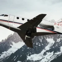 Ein weißer Businessjet der Finow Air Service mit der Kennung D-IETB befindet sich in einer steilen Steigflugphase. Das Flugzeug ist von schräg unten links vor der beeindruckenden Kulisse einer massiven, teilweise schneebedeckten Felswand eines Gebirges dargestellt. Der Rumpf zeigt die charakteristischen roten und blauen Zierlinien sowie das Firmenlogo am Heck. Die Aufnahme betont die Leistungsfähigkeit und Dynamik des Flugzeugs in alpiner Umgebung.