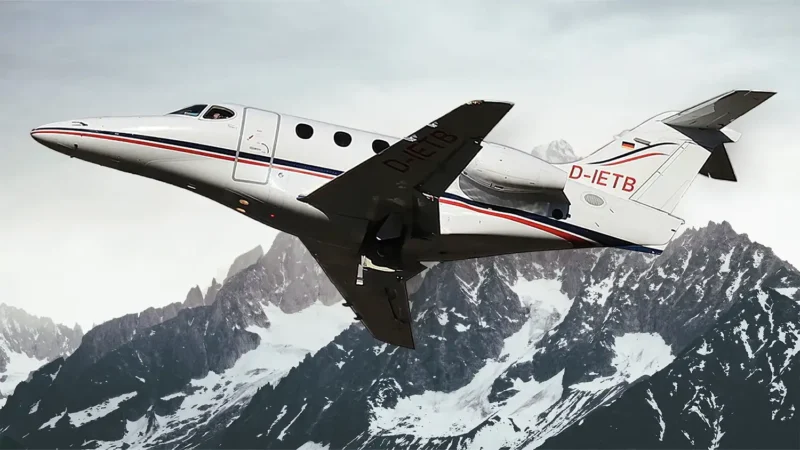 Ein weißer Businessjet der Finow Air Service mit der Kennung D-IETB befindet sich in einer steilen Steigflugphase. Das Flugzeug ist von schräg unten links vor der beeindruckenden Kulisse einer massiven, teilweise schneebedeckten Felswand eines Gebirges dargestellt. Der Rumpf zeigt die charakteristischen roten und blauen Zierlinien sowie das Firmenlogo am Heck. Die Aufnahme betont die Leistungsfähigkeit und Dynamik des Flugzeugs in alpiner Umgebung.