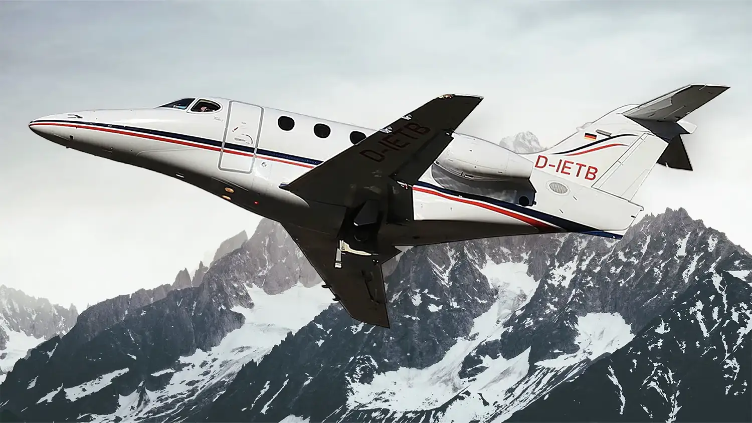 Ein weißer Businessjet der Finow Air Service mit der Kennung D-IETB befindet sich in einer steilen Steigflugphase. Das Flugzeug ist von schräg unten links vor der beeindruckenden Kulisse einer massiven, teilweise schneebedeckten Felswand eines Gebirges dargestellt. Der Rumpf zeigt die charakteristischen roten und blauen Zierlinien sowie das Firmenlogo am Heck. Die Aufnahme betont die Leistungsfähigkeit und Dynamik des Flugzeugs in alpiner Umgebung.