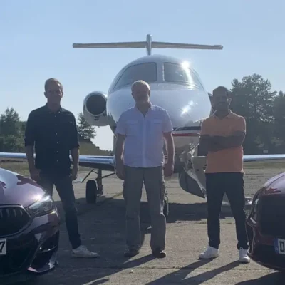 Drei Männer stehen in Frontalansicht auf einem sonnigen Rollfeld vor einem weißen Businessjet der Finow Air Service. Das Flugzeug ist zentral im Hintergrund positioniert. Im Vordergrund sind links und rechts die Frontpartien zweier dunkler Sportwagen (BMW) angeschnitten. Die Personen tragen Freizeitkleidung und blicken in die Kamera. Die Aufnahme vermittelt einen exklusiven Lifestyle-Charakter im Kontext von privater Luftfahrt und hochwertigen Fahrzeugen.