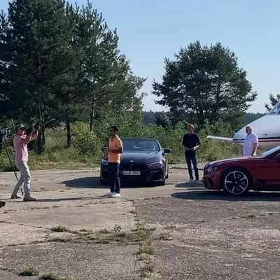 Eine Weitwinkelaufnahme zeigt eine Film- oder Videoserie-Produktion auf einem betonierten Rollfeld. Ein Kameramann links im Bild filmt einen Moderator in einem orangefarbenen Poloshirt, der vor einem dunkelblauen BMW Sportwagen steht. Im Hintergrund sind zwei weitere Männer sowie die Bugsektion eines weißen Businessjets der Finow Air Service vor einer Waldkulisse erkennbar. Rechts im Vordergrund ist zudem ein Teil eines roten Sportwagens (Bentley) zu sehen. Die Szene dokumentiert die Produktion von Lifestyle- oder Automobil-Content im Umfeld der privaten Luftfahrt.