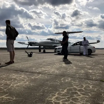 Eine Weitwinkelaufnahme auf einem betonierten Rollfeld zeigt die Ankunft oder Abfahrt von Passagieren bei dramatisch bewölktem Himmel. Im Zentrum steht ein zweimotoriges weißes Propellerflugzeug der Finow Air Service, neben dem eine weiße Mercedes-Benz Limousine mit geöffneter Fahrertür parkt. Drei Personen sind in der Szene verteilt: Ein Mann im Vordergrund links blickt zum Flugzeug, ein weiterer steht zentral am Pkw, und eine dritte Person in weißer Kleidung befindet sich rechts hinter dem Fahrzeug. Die Aufnahme vermittelt eine Atmosphäre von exklusivem Reiseservice und nahtloser Mobilität.