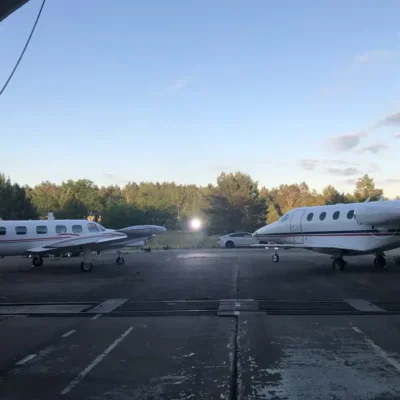 Eine Weitwinkelaufnahme zeigt zwei weiße Businessjets der Finow Air Service, die sich auf einem betonierten Vorfeld gegenüberstehen. Beide Flugzeuge tragen die charakteristischen roten und blauen Zierlinien. Im Hintergrund, zwischen den beiden Maschinen, ist ein heller Scheinwerfer einer Film- oder Fotoproduktion sowie ein geparkter Pkw vor einer Waldkulisse erkennbar. Die Szene ist bei beginnender Abenddämmerung unter einem klaren Himmel aufgenommen, was eine ruhige und professionelle Atmosphäre auf dem Flugplatzgelände vermittelt.