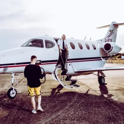 Ein Kameramann in gelben Shorts und schwarzem Langarmshirt filmt mit einem Gimbal-Stabilisierungssystem einen Piloten, der gerade über die integrierte Treppe aus einem weißen Businessjet der Finow Air Service aussteigt. Der Pilot trägt eine weiße Uniform mit Krawatte. Das Flugzeug mit der Kennung D-IFTR steht auf einem betonierten Rollfeld bei hellem Tageslicht. Die Szene zeigt eine professionelle Videoaufnahme für Werbe- oder Dokumentationszwecke von Finow Air Service.