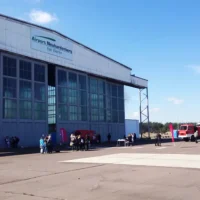 Eine Außenaufnahme zeigt einen großen Flugzeughangar auf dem Gelände des Airport Neuhardenberg. An der oberen Fassade des Hangars ist das Logo des Flughafens mit dem Schriftzug „Airport Neuhardenberg bei Berlin“ angebracht. Die großen Hangartore aus Metall und Glas sind teilweise geschlossen. Auf dem Vorfeld vor dem Hangar befinden sich mehrere Personengruppen, kleine Infostände und ein rotes Feuerwehrfahrzeug. Die Szene ist bei sonnigem Wetter unter einem blauen Himmel aufgenommen und dokumentiert eine Veranstaltung oder einen Besuchertag auf dem Flugplatzgelände.