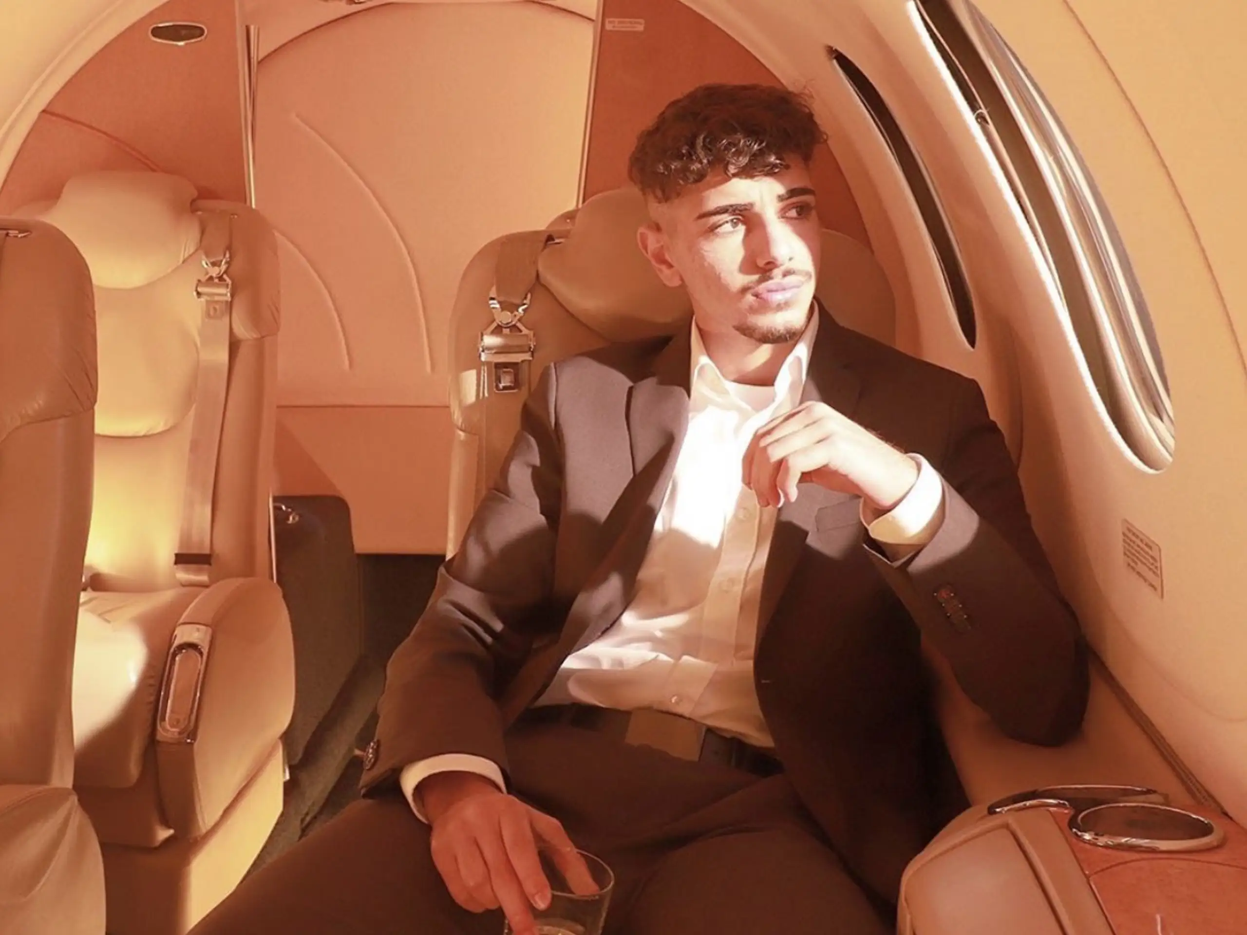Ein junger Mann in einem dunklen Anzug und weißem Hemd sitzt entspannt in einem der beigefarbenen Ledersitze in der Kabine eines Businessjets. Er blickt aus dem Fenster auf der rechten Seite, während helles Sonnenlicht in die Kabine fällt. In seiner rechten Hand hält er ein Glas. Die Kabinenausstattung ist in warmen Beigetönen gehalten, und im Hintergrund sind weitere Passagiersitze sowie die Rückwand der Kabine erkennbar. Die Szene vermittelt eine ruhige und exklusive Reiseatmosphäre an Bord eines Flugzeugs von Finow Air Service.
