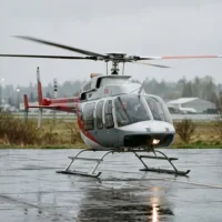 Ein grau-rot lackierter Hubschrauber steht mit laufenden Rotoren auf einem nassen, spiegelnden Asphaltboden eines Flugplatzes. In der Cockpitkanzel sind zwei Personen erkennbar. Der Hubschrauber ist in Frontalansicht dargestellt, wobei die Landekufen und das eingeschaltete Buglicht deutlich sichtbar sind. Der Hintergrund zeigt eine neblige, herbstliche Landschaft mit kahlen Bäumen und einem Flughafengebäude unter einem grauen, bedeckten Himmel. Die Aufnahme verdeutlicht die Allwetter-Einsatzbereitschaft und die Erweiterung des Portfolios von Finow Air Service um Drehflügler.