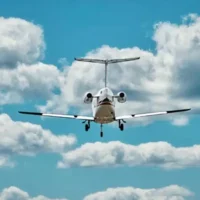 Ein weißer Businessjet der Finow Air Service ist von hinten im direkten Anflug dargestellt. Das Fahrwerk ist vollständig ausgefahren. Das Flugzeug befindet sich zentral im Bild vor einem blauen Himmel mit markanten, weißen Quellwolken. Die Triebwerke an den Seiten des Hecks sowie das T-Leitwerk sind aus dieser Perspektive deutlich erkennbar. Die Aufnahme vermittelt die Dynamik des Landeanflugs.