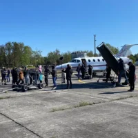 Eine Weitwinkelaufnahme zeigt ein Filmset auf einem weitläufigen, betonierten Flugplatzgelände unter klarem blauen Himmel. Im Zentrum steht eine große Filmcrew mit technischem Equipment, darunter ein Kamerakran (Dolly) und Mikrofone. Im Hintergrund ist ein weißer Businessjet der Finow Air Service mit der Kennung D-IETB positioniert, an dem eine schwarze Abschirmung oder Lichtblende aufgestellt wurde. Die Szene verdeutlicht die Nutzung von Finow Air Service Flugzeugen als Filmrequisite oder Drehort für Medienproduktionen. Waldränder und Flughafengebäude begrenzen das Areal im Hintergrund.