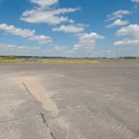 Eine Weitwinkelaufnahme zeigt eine große, betonierte Vorfeldfläche eines Flugplatzes unter einem blauen Himmel mit lockeren weißen Wolken. Die Betonplatten weisen stellenweise Ausbesserungen und Sandablagerungen auf. Im Hintergrund erstreckt sich eine weite Graslandschaft, hinter der eine großflächige Photovoltaikanlage (Solarpark) erkennbar ist. Vereinzelt stehen Lichtmasten am Rand des Geländes. Die Szene vermittelt die Weitläufigkeit und die technische Infrastruktur des Flugplatzes.