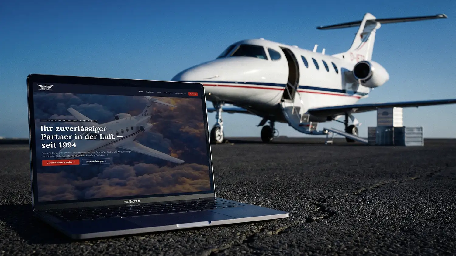 Eine Außenaufnahme auf einem asphaltierten Rollfeld bei sonnigem Wetter. Im Vordergrund steht ein geöffneter Laptop (MacBook Pro), auf dessen Bildschirm die Startseite der Finow Air Service Website mit dem Slogan „Ihr zuverlässiger Partner in der Luft – seit 1994“ zu sehen ist. Im Hintergrund parkt ein weißer Businessjet der Finow Air Service mit der Kennung D-IETB, dessen Passagiertür geöffnet ist. Die Szene kombiniert die digitale Präsenz des Unternehmens mit dem realen Flugbetrieb auf dem Flugplatz.