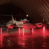 Eine Weitwinkelaufnahme zeigt ein professionelles Fotoshooting in einem großen, bogenförmigen Flugzeughangar. Ein weißer Businessjet der Finow Air Service ist im Hintergrund positioniert und wird durch vertikale, rot leuchtende Lichtstäbe sowie helle Spotlichter und künstlichen Nebel inszeniert. Vor dem Flugzeug steht eine Person in heller Kleidung. Im Vordergrund sind technisches Equipment, darunter ein Stativ mit einem großen schwarzen Lichtformer (Softbox) und Kabel am Boden, erkennbar. Die gesamte Szene ist in ein intensives rotes Licht getaucht, was eine moderne und exklusive Bildwirkung erzeugt.