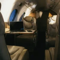 Eine Innenaufnahme der Passagierkabine eines Businessjets von Finow Air Service. Die Kabine ist mit beigefarbenen Ledersitzen in einer Club-Anordnung ausgestattet. Auf einem ausklappbaren Holztisch im Vordergrund steht ein geöffneter Laptop neben einem Smartphone. Das warme, punktuelle Deckenlicht beleuchtet die Sitzgruppe und schafft eine ruhige Arbeitsatmosphäre. Durch die ovalen Kabinenfenster fällt diffuses Tageslicht. Die Szene verdeutlicht die Nutzung des Flugzeugs als mobiles Büro während des Reiseflugs.