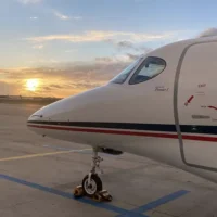 Eine Nahaufnahme der Bugsektion eines weißen Businessjets vom Typ Beechcraft Premier I der Finow Air Service. Das Flugzeug steht auf einem betonfreien Vorfeld und ist mit gelben Bremsklötzen am Bugrad gesichert. Der Rumpf zeigt die charakteristischen roten und blauen Zierstreifen sowie technische Markierungen an der Kabinentür. Im Hintergrund ist ein stimmungsvoller Sonnenuntergang über dem Flugplatzgelände zu sehen, wobei die Sonne tief am Horizont hinter einer leichten Wolkendecke steht. Im fernen Hintergrund sind die Silhouetten weiterer geparkter Flugzeuge erkennbar.