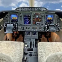 Eine detaillierte Innenaufnahme des Cockpits eines modernen Businessjets. Das Instrumentenpanel umfasst drei große digitale Multifunktionsdisplays (Glass Cockpit), die Flugdaten, Navigationskarten und Systeminformationen anzeigen. Davor befinden sich die beiden Steuerhörner für Pilot und Co-Pilot. Die Sitze sind mit hellen Lammfellbezügen ausgestattet. In der Mittelkonsole sind Schubhebel, Tastaturen für das Flight Management System sowie zahlreiche Schalter und Regler für die Flugzeugsteuerung angeordnet. Durch die Cockpitfenster ist ein blauer Himmel mit weißen Wolken erkennbar.
