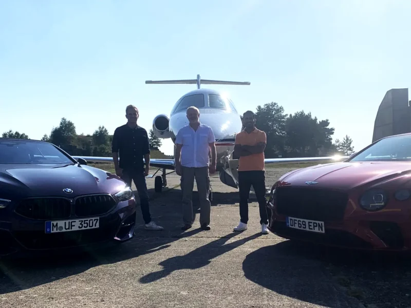 Drei Männer stehen in Frontalansicht auf einem sonnigen Rollfeld vor einem weißen Businessjet der Finow Air Service. Das Flugzeug ist zentral im Hintergrund positioniert. Im Vordergrund sind links und rechts die Frontpartien zweier dunkler Sportwagen (BMW) angeschnitten. Die Personen tragen Freizeitkleidung und blicken in die Kamera. Die Aufnahme vermittelt einen exklusiven Lifestyle-Charakter im Kontext von privater Luftfahrt und hochwertigen Fahrzeugen.