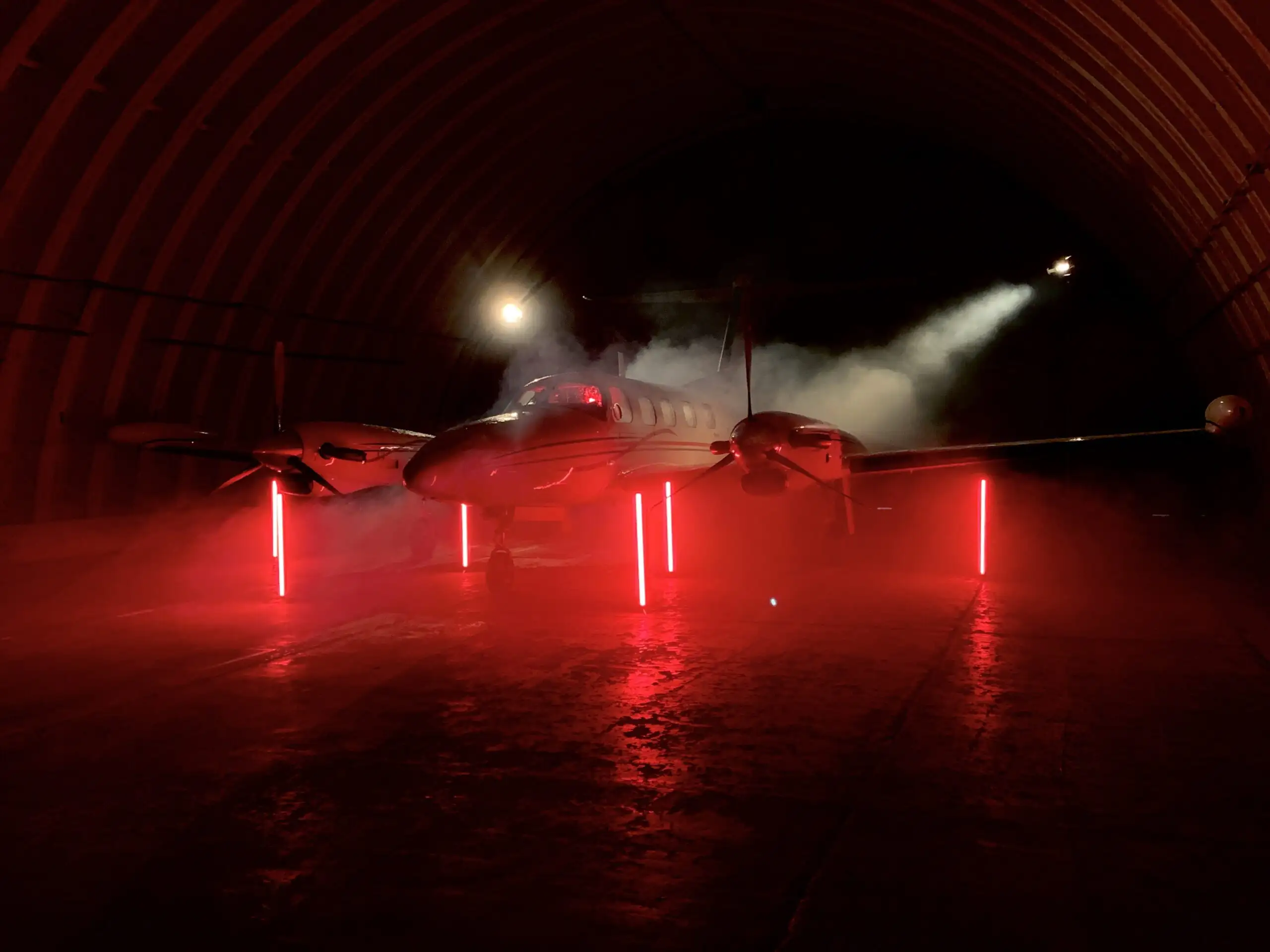 Eine künstlerische und dramatisch beleuchtete Aufnahme eines Businessjets der Finow Air Service in einem dunklen, bogenförmigen Hangar. Das Flugzeug ist von vorne fotografiert und wird von mehreren vertikalen, rot leuchtenden Lichtstäben am Boden sowie einem hellen weißen Scheinwerferstrahl von oben rechts inszeniert. Künstlicher Nebel umspült den Rumpf und die Tragflächen, was zusammen mit den roten Lichtreflexionen auf dem glänzenden Boden eine mystische und exklusive Atmosphäre schafft. Die Szene dokumentiert eine professionelle Foto- oder Videoproduktion zur Markeninszenierung.