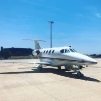 Ein weißer Businessjet der Finow Air Service mit der Kennung D-IETB steht auf einem betonierten Vorfeld unter strahlend blauem Himmel. Das Flugzeug ist von schräg vorne rechts fotografiert und zeigt die charakteristischen roten und blauen Zierlinien am Rumpf. Im Hintergrund sind ein hoher Beleuchtungsmast und Flughafengebäude erkennbar. Die Szene wirkt hell und sonnig.