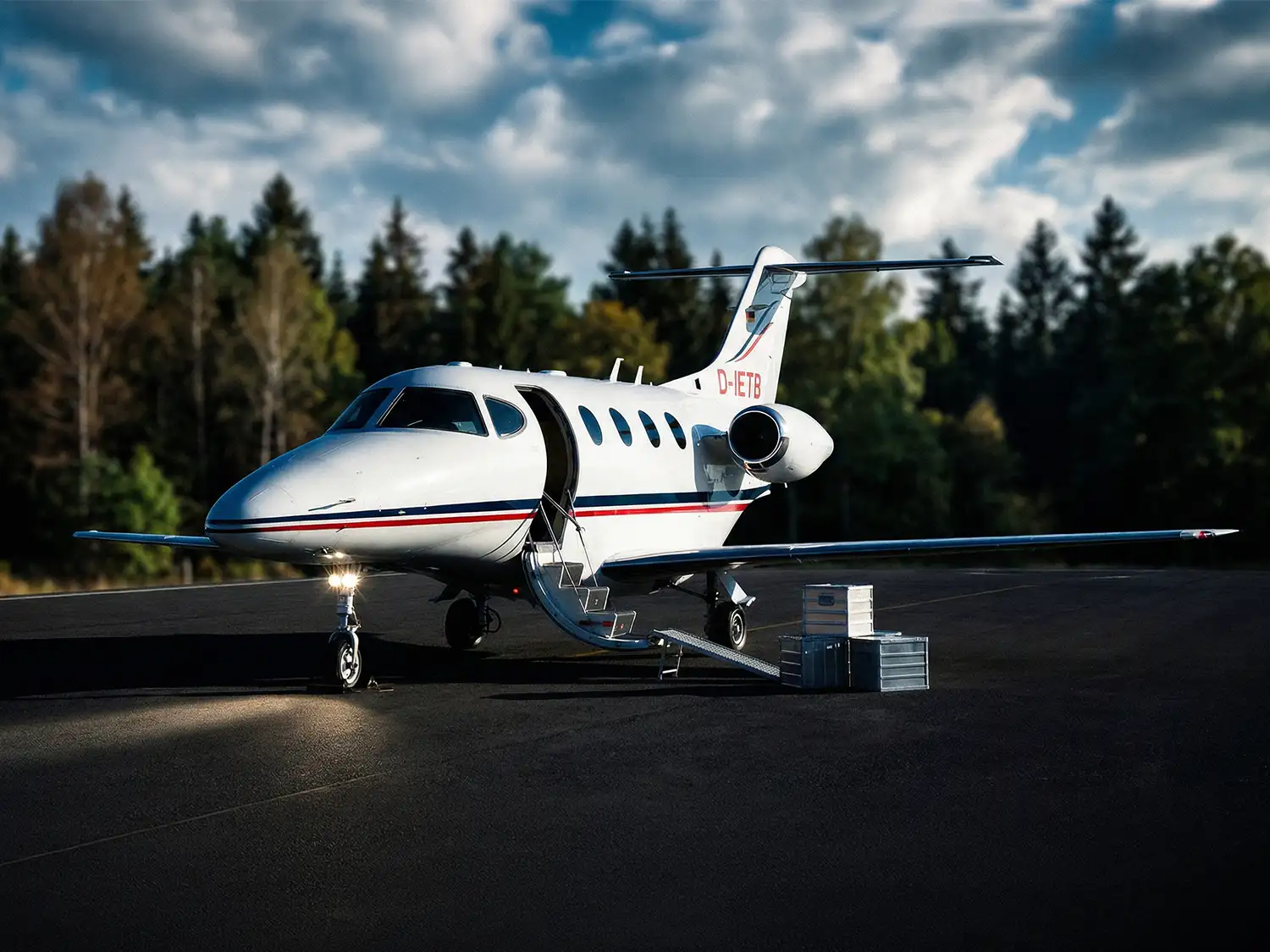 Ein weißer Businessjet der Finow Air Service mit der Kennung D-IETB steht auf einem dunklen Rollfeld vor einer Waldkulisse unter einem dramatisch bewölkten Himmel. Die Passagiertür ist geöffnet und die integrierte Treppe ausgefahren. Daneben ist eine Rampe am Boden platziert, über die mehrere silberne Aluminium-Transportkisten verladen werden. Das Buglicht des Flugzeugs ist eingeschaltet und wirft einen hellen Lichtkegel auf den Asphalt. Die Szene verdeutlicht die Nutzung des Flugzeugs für den kombinierten Transport von Passagieren und hochwertiger Fracht.