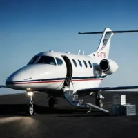 Ein weißer Businessjet der Finow Air Service mit der Kennung D-IETB steht auf einem dunklen Rollfeld vor einer neutral blauen Kulisse. Die Passagiertür ist geöffnet und die integrierte Treppe ausgefahren. Daneben ist eine Rampe am Boden platziert, über die mehrere silberne Aluminium-Transportkisten verladen werden. Das Buglicht des Flugzeugs ist eingeschaltet und wirft einen hellen Lichtkegel auf den Asphalt. Die Szene verdeutlicht die Nutzung des Flugzeugs für den kombinierten Transport von Passagieren und hochwertiger Fracht.